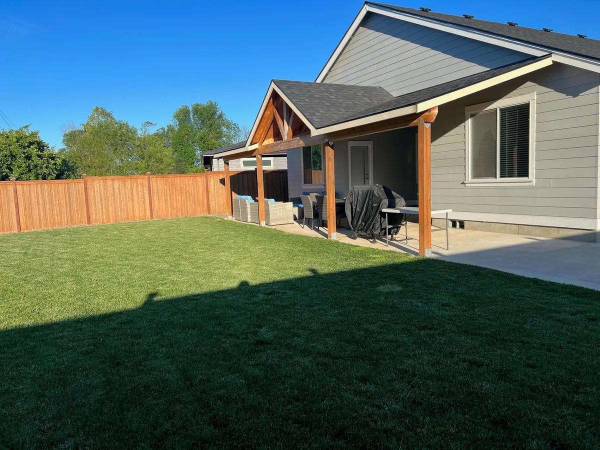 This image showcases a spacious backyard with a lush green lawn, bordered by a tall wooden fence for complete privacy and safety. A covered patio area extends from the home, furnished with comfortable outdoor seating and a table, perfect for gatherings. The overall setup creates a relaxed, family-friendly environment ideal for play, dining, and enjoying the outdoors.