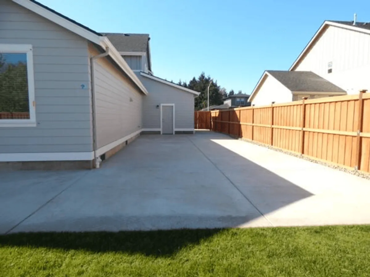 A bright, inviting backyard with a concrete patio, lush green grass, and a secure wooden fence. The scene shows ample space for kids to play, a safe environment for family activities, and plenty of room for outdoor gatherings such as barbecues or birthday parties.