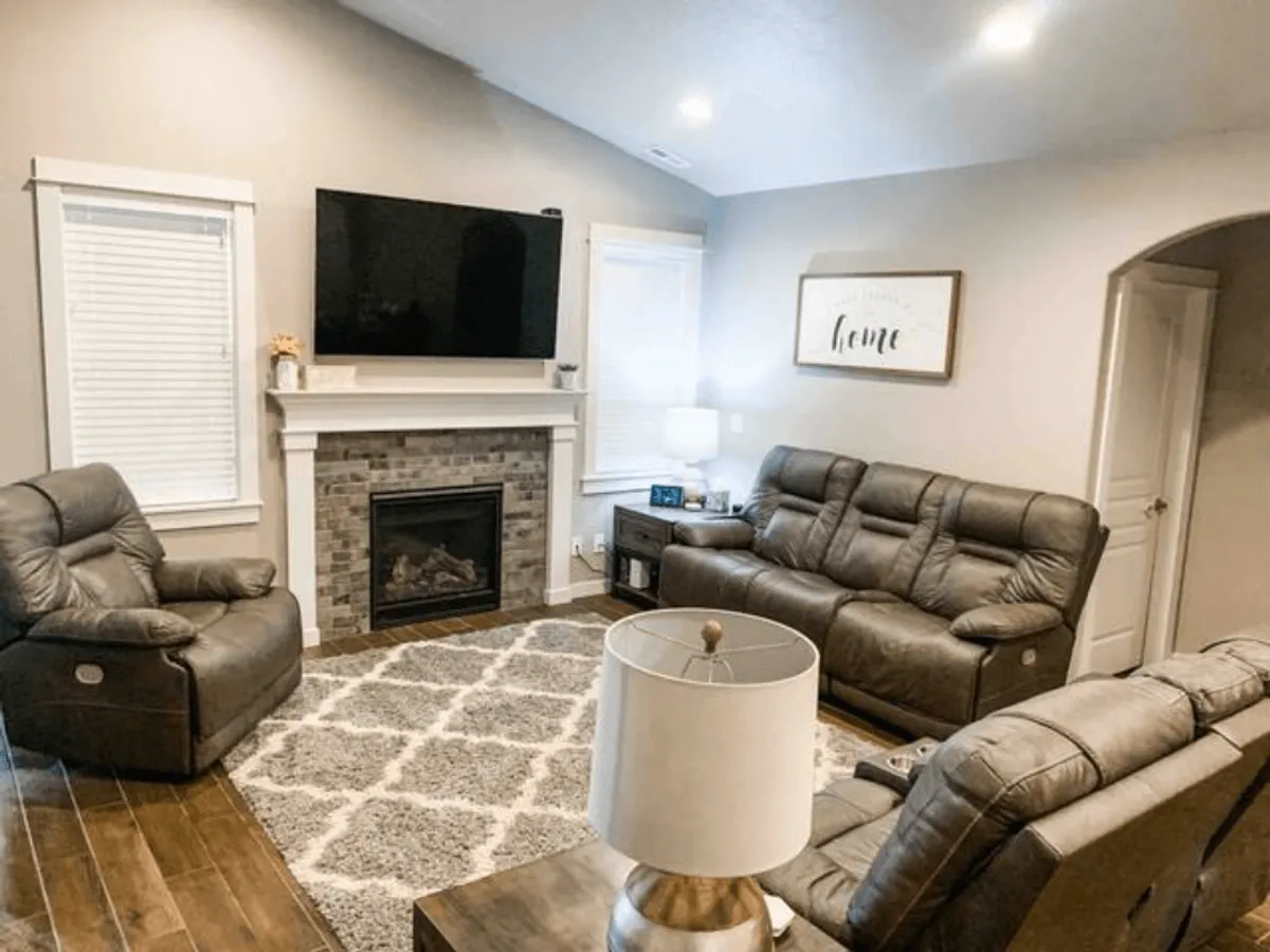 A warm, inviting living room featuring high vaulted ceilings, a modern fireplace, plush electric recliners, a smart TV mounted above the mantle, and a large area rug. The space is bathed in natural light, with ample seating for groups to gather and relax.
