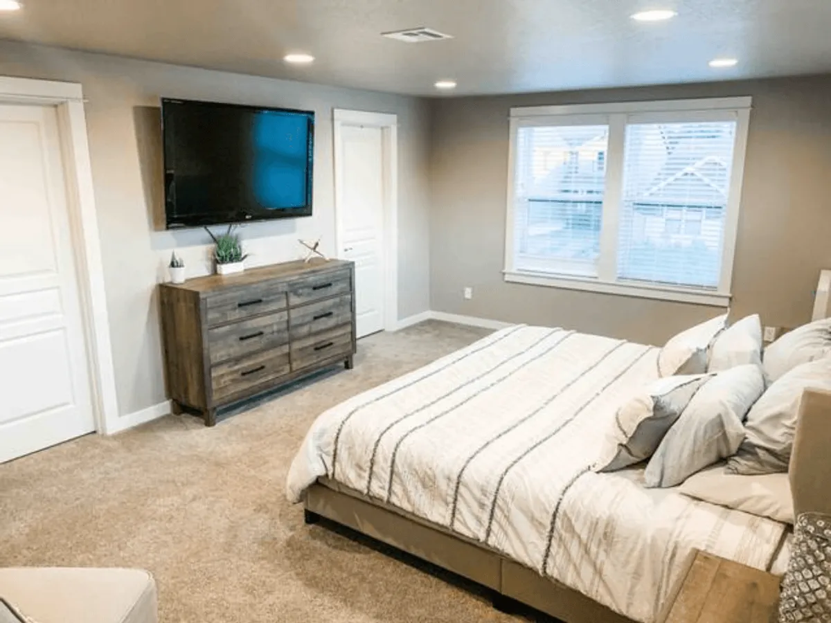This tranquil bedroom features a stylish bed with striped bedding, a solid wooden dresser, a flat-screen TV, and windows letting in bright, natural light. The setting suggests a peaceful night's sleep and modern comfort.
