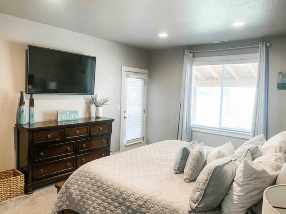 A cozy bedroom is presented with a well-made bed, decorative pillows, a dresser topped with a flat-screen smart TV, and a large window bringing in abundant natural light. The room exudes comfort and a sense of personal retreat.