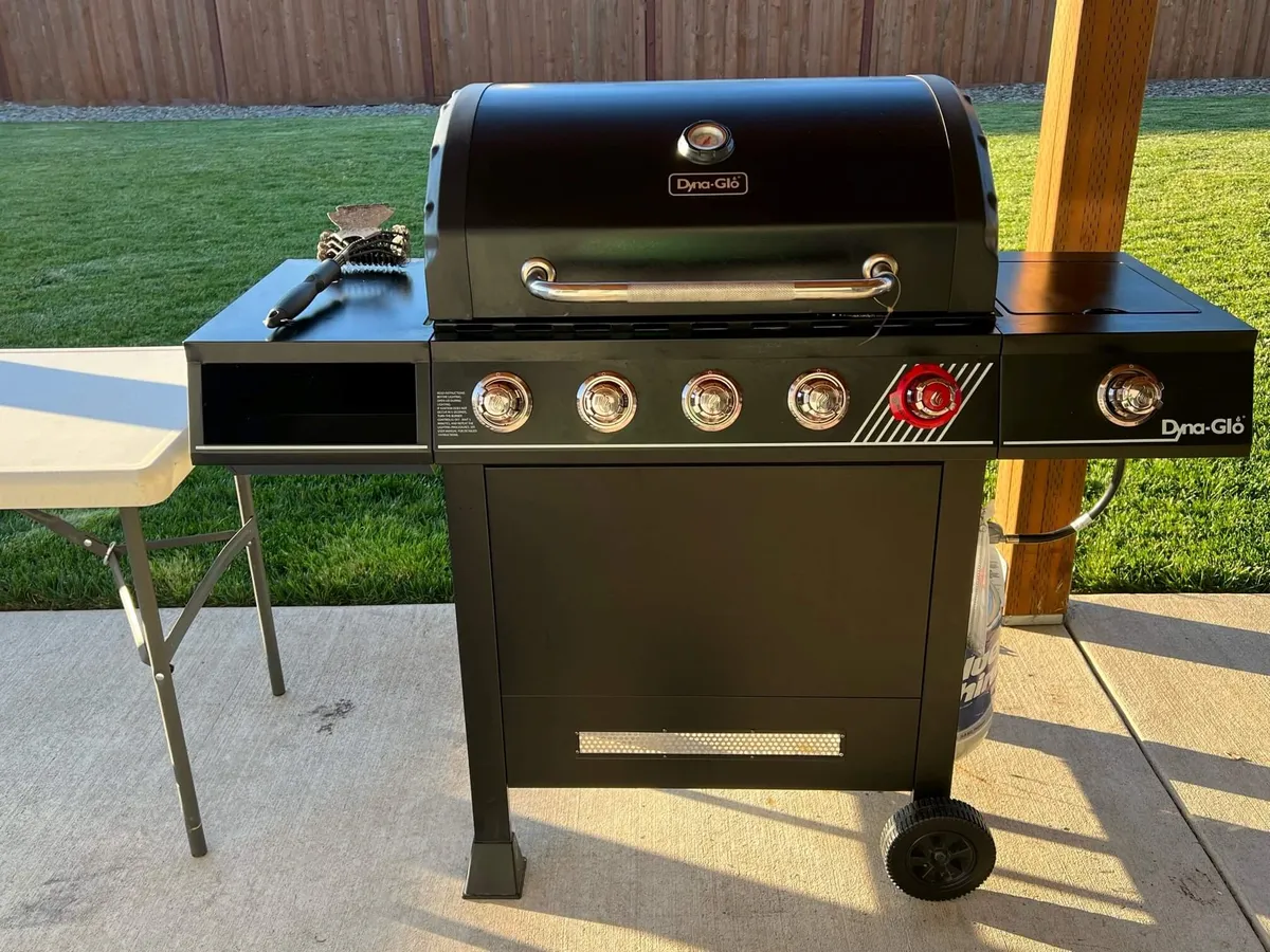 A close-up of an outdoor grill and side table, situated on a patio near a grass lawn and privacy fence. The setup suggests a casual, festive atmosphere ideal for cookouts and socializing with friends or family.